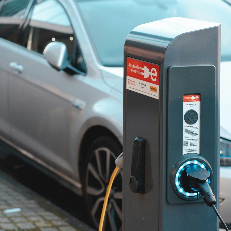 Ladesäule für Elektroautos mit angeschlossenem Ladekabel und einem grauen Auto im Hintergrund.